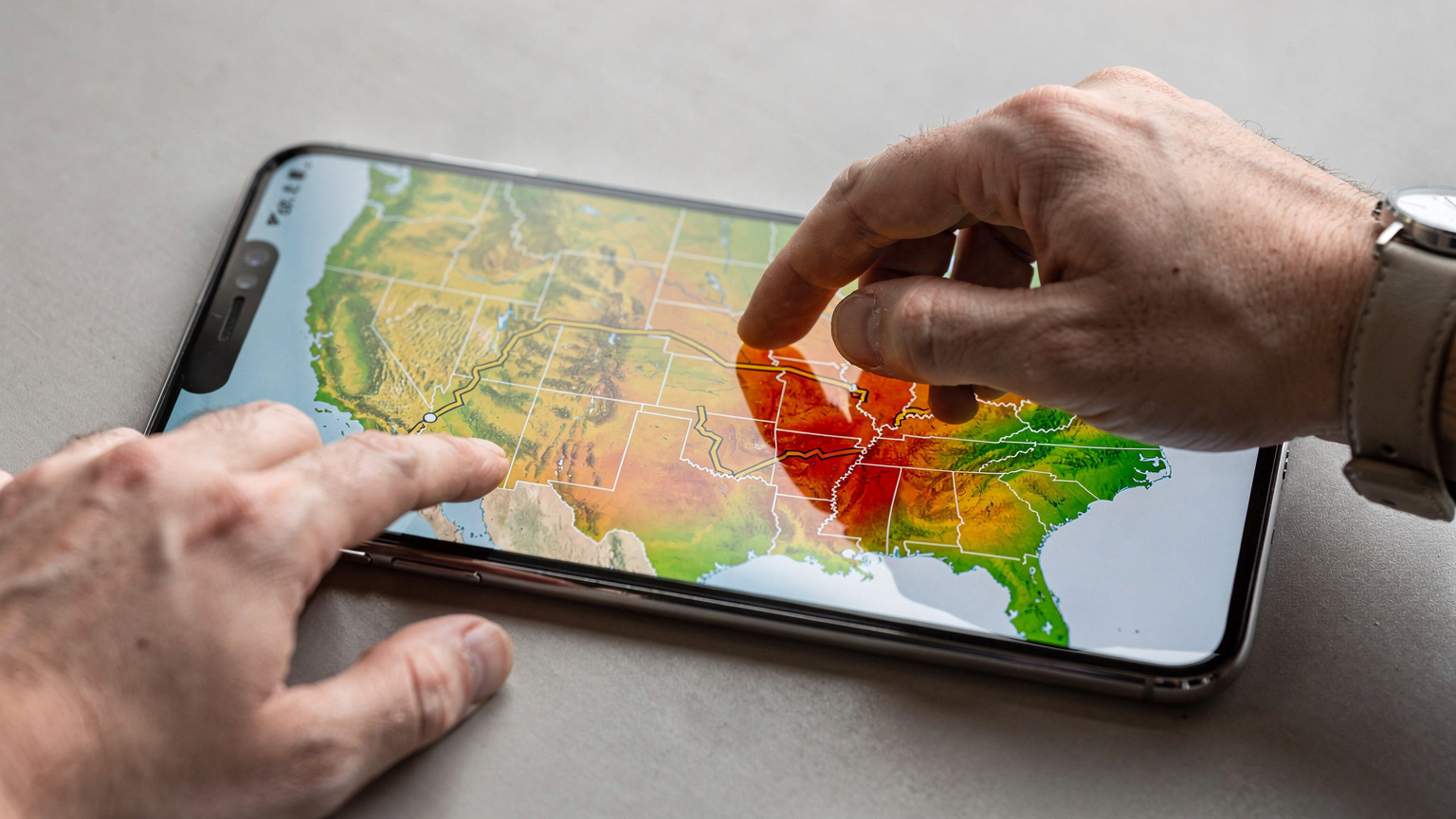 A close-up of a digital map displayed on a smartphone, showing a winding cross-country road trip route with different colored overlays indicating varying levels of mobile network coverage, from strong to weak. A traveler's finger is tracing a section of the route.