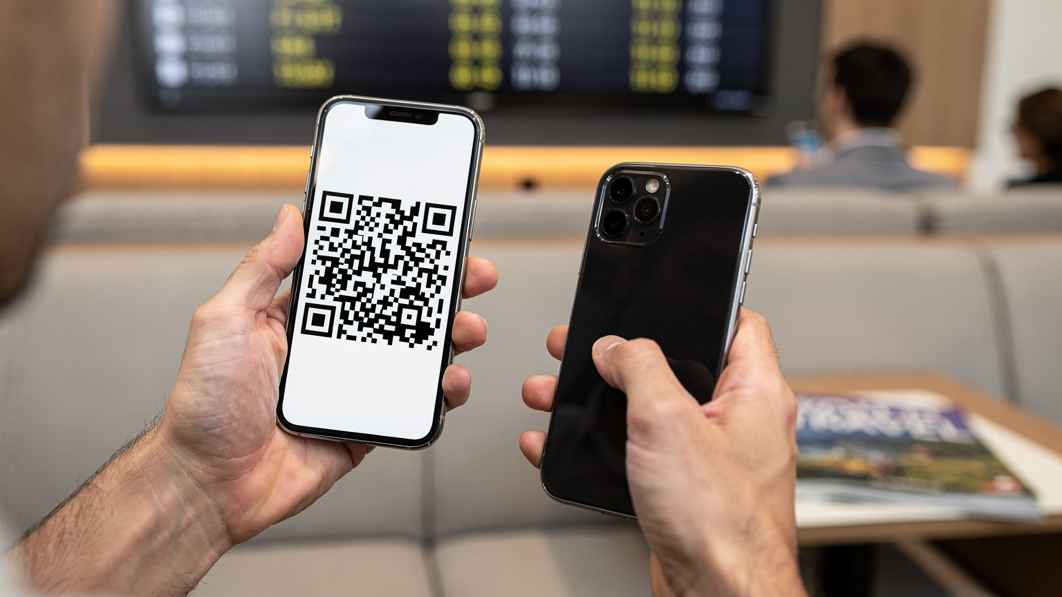 Close-up of human hands, one holding a smartphone displaying a QR code, the other holding a second phone scanning it, at an airport lounge.