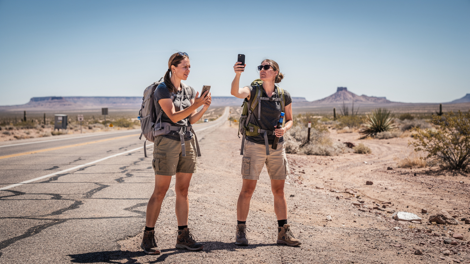 Una viajera solitaria, de perfil, mirando atentamente su smartphone en un paisaje desértico con colinas bajas y cielos azules, buscando la mejor conexión posible para su ruta.