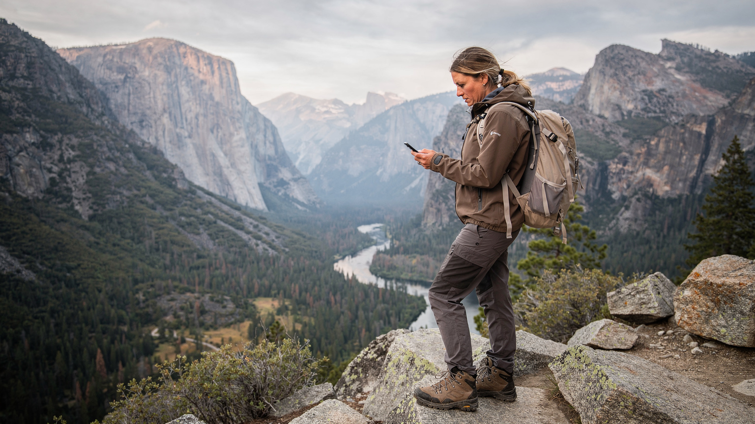 A woman stands at a remote national park overlook, looking at her phone with a concerned expression, against a backdrop of vast forests and distant mountains under a dramatic sky.