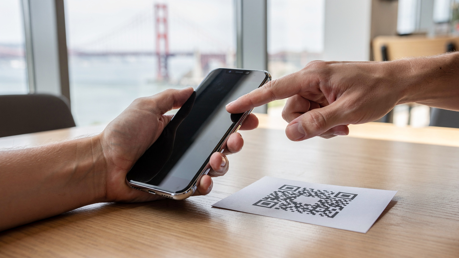 Due mani tengono uno smartphone, mentre un dito tocca delicatamente lo schermo per scansionare un codice QR su un foglio di carta, su un tavolo di un caffè con vista sul Golden Gate Bridge.