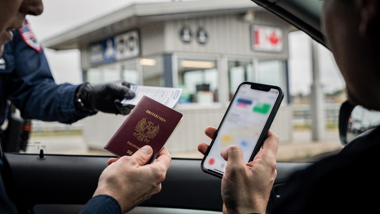 Ein Reisender in einem Auto reicht an einem Landgrenzübergang zwischen den USA und Kanada seinen Reisepass einem Grenzbeamten, während er sein Smartphone mit einer geöffneten App in der anderen Hand hält.