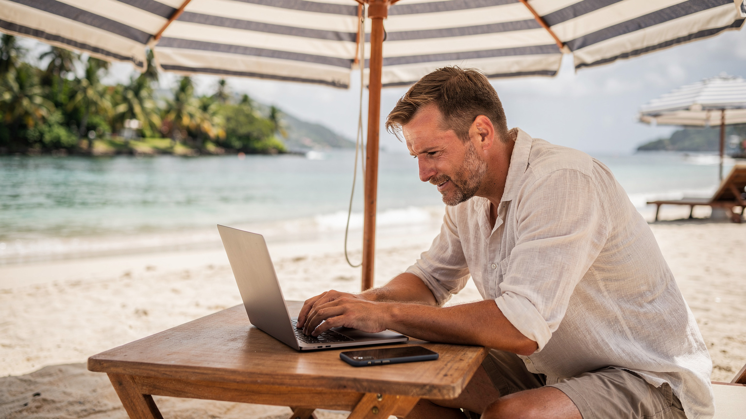 Un homme travaillant sur un ordinateur portable, son smartphone à côté, sous un parasol sur une plage des Caraïbes, avec l'océan turquoise et des palmiers en arrière-plan. Il est serein et concentré.