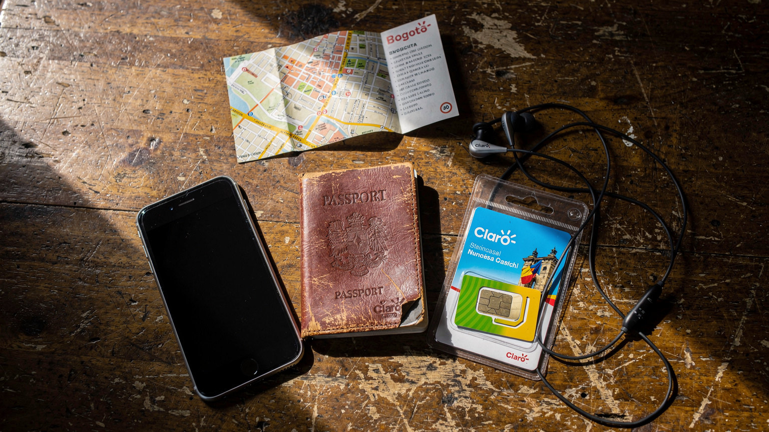 An overhead flat-lay shot on a rustic wooden table in a Colombian hostel, featuring a passport, a smartphone, a local SIM card package, and a small, well-worn map of Bogotá, indicating a traveler's preparation and backup plans.