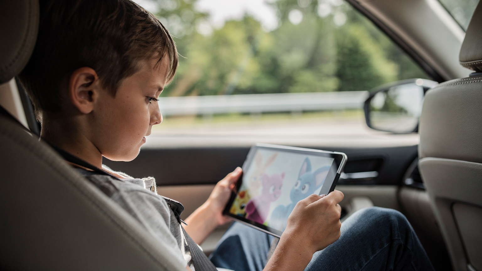 A child is engrossed in watching an animated movie on an iPad in the backseat of a car, with the reflection of a highway and passing trees visible on the screen, indicating a long journey.