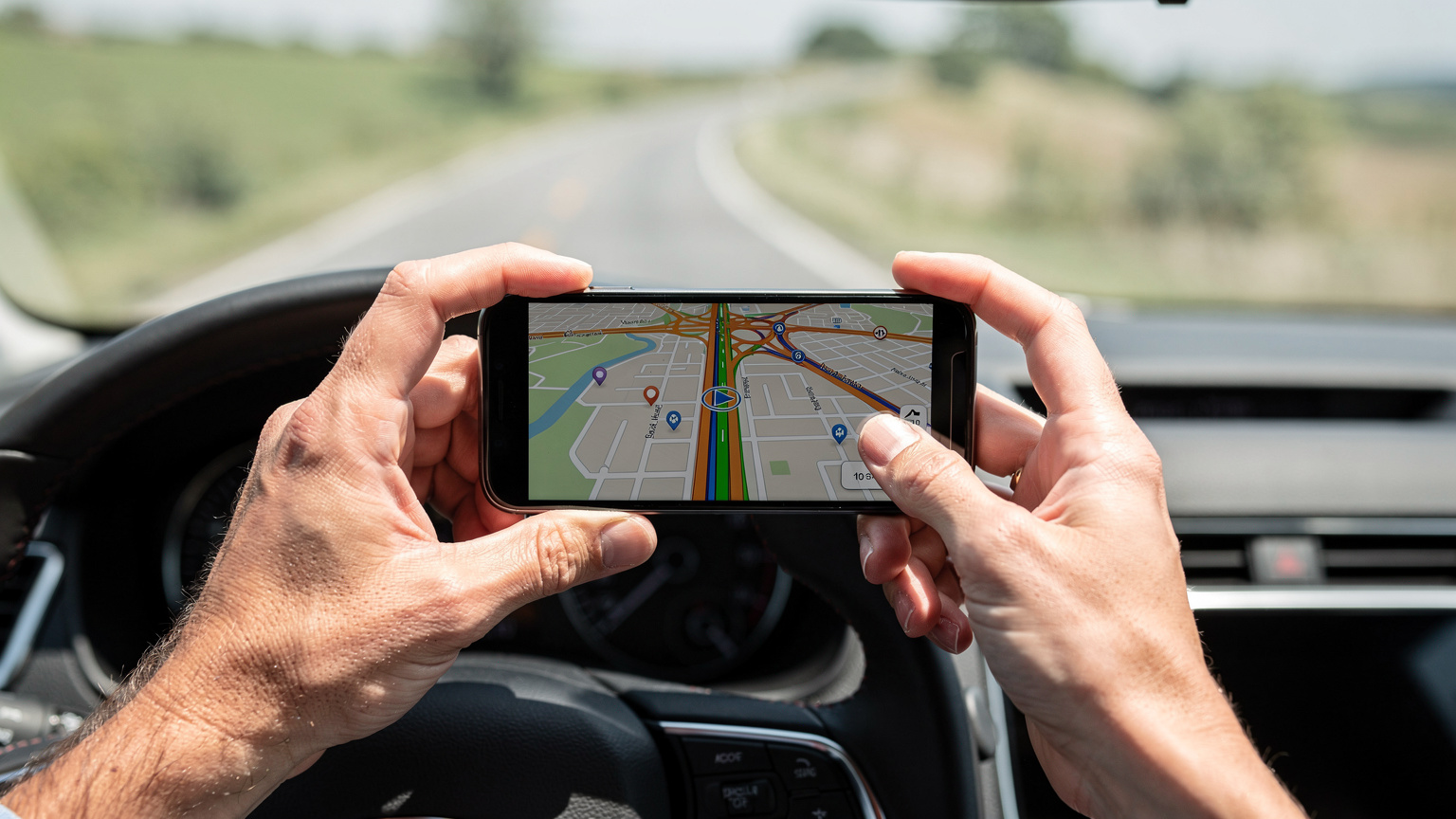 Gros plan sur les mains d'un voyageur tenant un smartphone, montrant un itinéraire GPS détaillé sur l'écran. La lumière du soleil éclaire l'habitacle de la voiture, et l'arrière-plan flou suggère une route ouverte traversant des champs.