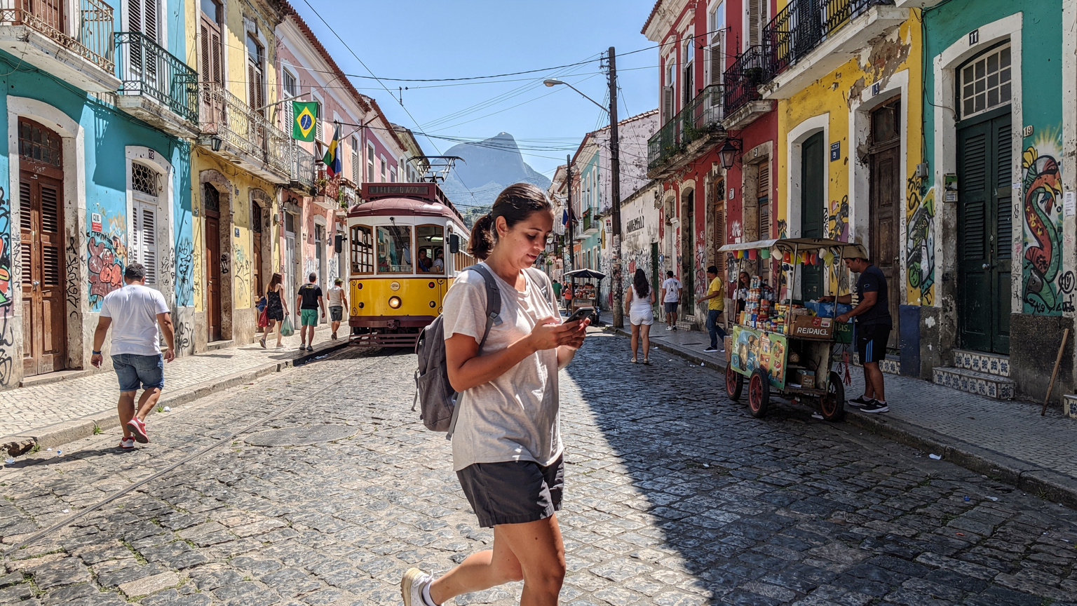 Uma turista caminhando por uma rua movimentada no Centro Histórico do Rio de Janeiro, com prédios coloniais coloridos e bondes ao fundo, enquanto consulta um mapa em seu Google Pixel 9.